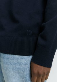 Close-up van een hand die een marineblauwe trui met een geribde zoom draagt, gecombineerd met lichtblauwe denim jeans. Subtiel logo zichtbaar op de trui.