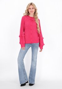 Cardigan fucsia con maniche arricciate e colletto, dotato di bottoni circolari e maglia texturizzata. Abbinato a jeans blu svasati e stivali neri.