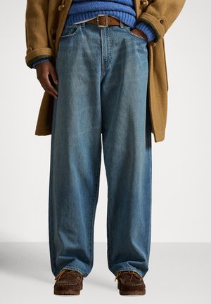 Φαρδιά τζιν παντελόνια από γαλάζιο ντένιμ συνδυασμένα με καφέ ζώνη και μοκασίνια σκούρου καφέ χρώματος, φορεμένα κάτω από ένα καμηλό παλτό.