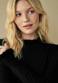 Maglione nero a coste con scollatura e polsini arricciati, indossato con uno stile casual. La modella ha capelli biondi ondulati e trucco naturale.