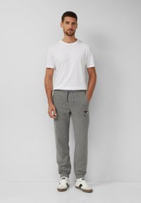 Man in een wit T-shirt met korte mouwen, grijze sweatpants met een Snoopypatch en witte sneakers met zwarte accenten, leunend tegen een witte muur.