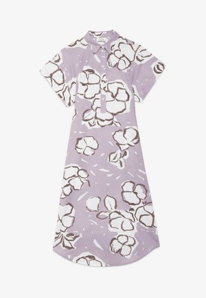 Mujer con vestido lila claro hasta la rodilla con estampados florales blancos y marrones, mangas cortas y zapatos planos beige, de pie con las manos en los bolsillos.