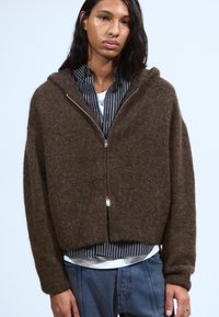 Haut zippé marron en matière texturée, avec un design court et des manches longues, porté par-dessus une chemise rayée.