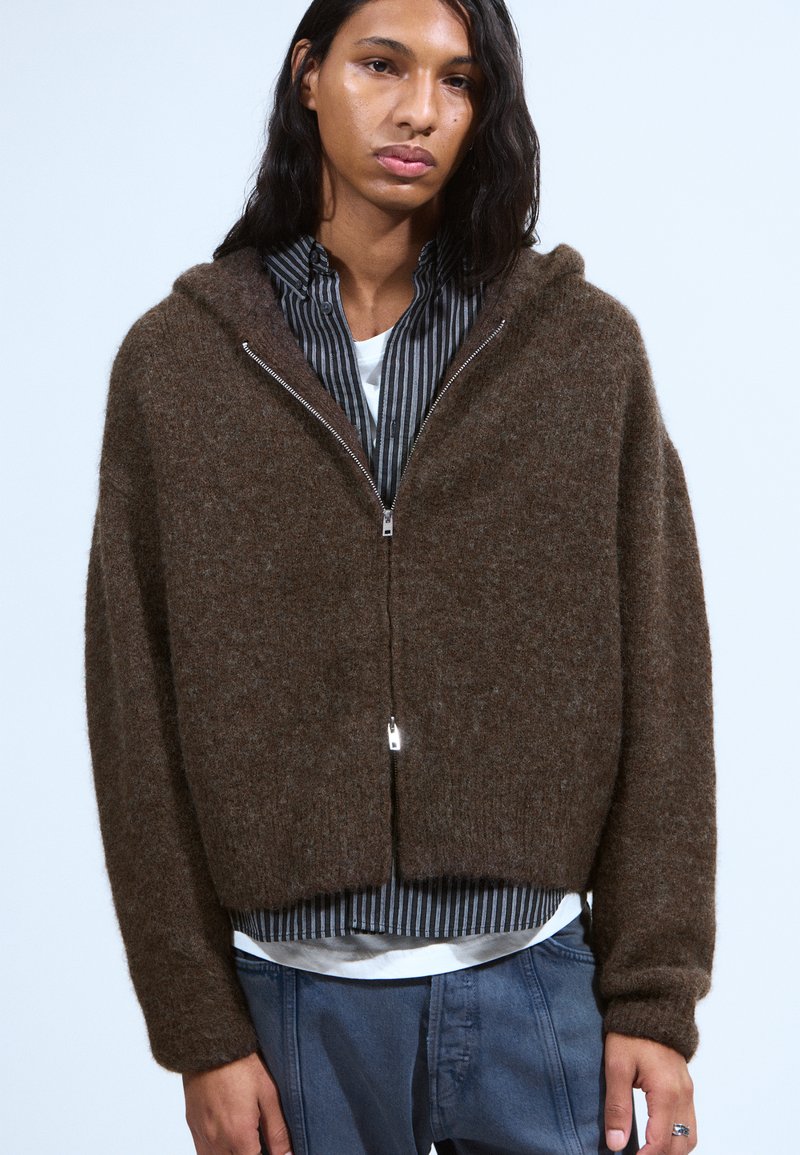 Haut zippé marron en matière texturée, avec un design court et des manches longues, porté par-dessus une chemise rayée.