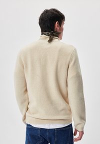 Beige Strickpullover mit hohem Kragen, geripptem Saum und Bündchen, getragen über einem weißen Hemd. Die Rückseite zeigt eine glatte Textur und eine entspannte Passform.