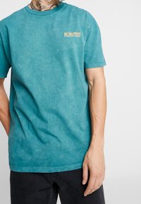 Camiseta de algodón turquesa con un aspecto desgastado, que presenta "BUDAPEST" en naranja en el lado izquierdo del pecho. Manga corta y cuello redondo.