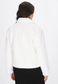 Femeie cu păr lung și închis la culoare, îmbrăcată într-o geacă de blană artificială albă și fustă neagră, surprinsă din spate pe un fond alb simplu.