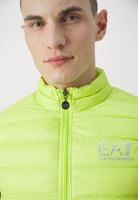 Svijetlo neonsko zelena podstavljena jakna s visokim ovratnikom, zatvaranjem na zip i EA7 logotipom na prsima. Ima kvadrantnu teksturu i glatki materijal.