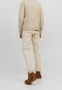 Une personne portant un pull beige, un pantalon crème et des bottes en suède marron, se tenant de dos contre un fond clair et uni.