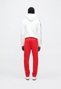 Bílá mikina s kapucí a černými pruhy po stranách, doplněná červenými tepláky s bílými bočními akcenty, pohled zezadu.