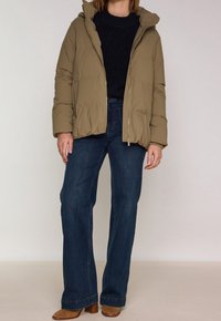 Žena ve světlém pufovaném kabátu, tmavě modrém svetru, širokých modrých džínách a béžových semišových kozačkách na podpatku stojí na jednobarevném pozadí.