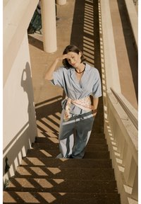 Camicia azzurro chiaro, pantaloni larghi con una fascia decorata da fiori; in piedi su una scala con ombre proiettate dalla ringhiera.