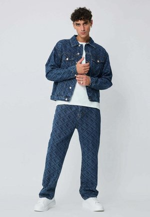 Jeune homme portant une veste en jean bleu foncé à motifs assortis et un pantalon avec des baskets blanches, debout devant un fond clair uni.