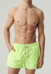 Neonowe zielone kąpielówki wykonane z lekkiego materiału, z elastycznym paskiem w talii, zaciągnięciem na sznurek oraz bocznymi kieszeniami.