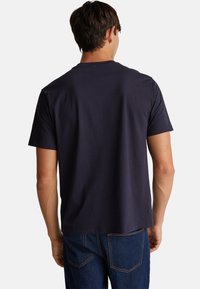 Mężczyzna w gładkiej, ciemnoniebieskiej koszulce z krótkim rękawem i niebieskich dżinsach, odwrócony plecami w stronę jednolitego, białego tła.