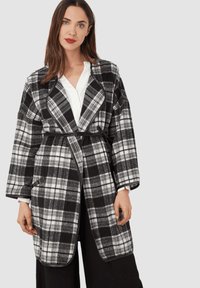 Abrigo envolvente a cuadros en blanco y negro con mangas largas, un ajuste holgado, bolsillos laterales y un cinturón de tela. Camisa blanca debajo.