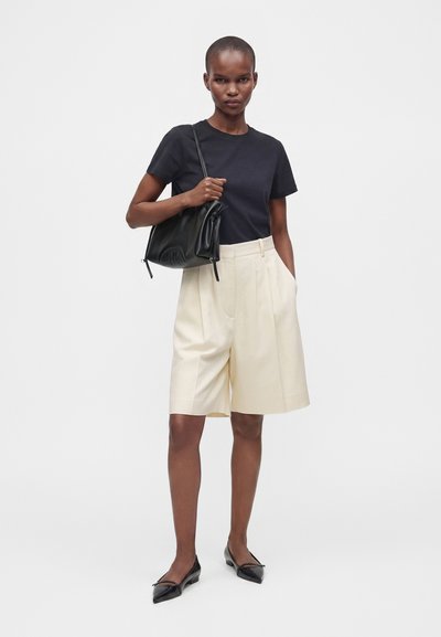 Femme portant un t-shirt noir et un short large crème longueur genoux, tenant un sac à main noir sur une épaule, debout devant un fond uni.