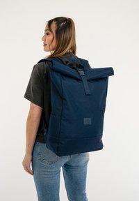 Zaino roll-top navy realizzato in tessuto resistente. Presenta spallacci regolabili, una tasca frontale e una patch con logo. Texture liscia e design minimalista.