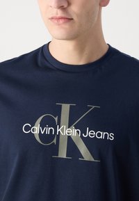 Camiseta de algodón azul marino con cuello redondo. Presenta un gran texto gráfico en contraste "Calvin Klein Jeans" con un destacado logo "CK".
