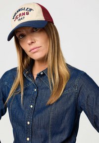 Puuvillane pesapalli müts, mille eesmine osa on beež, külgede võrkpaneelid on marooni värvi ja visor on tumesinine. Omab borduuritud "Wrangler Jeans 1947" teksti.