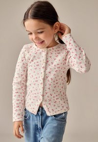 Bílý žebrovaný cardigan s malými růžovými květinovými vzory, dlouhé rukávy, kulatý výstřih a zapínání na knoflíky, v kombinaci s modrými denimovými džíny.