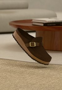Καφέ σκούρο παπούτσι τύπου slip-on με χρυσή αγκράφα και υφή στη σόλα, αιωρούμενο πάνω από μπεζ χαλί σε ένα σαλόνι με ουδέτερους τόνους.