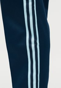 Tamnoplave hlače s tri okomite svijetloplave i crne pruge koje se spuštaju niz bočnu stranu nogavice na jednobojnoj pozadini.