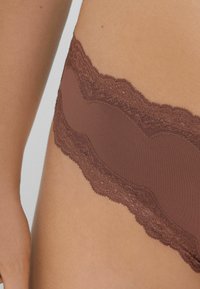 Gros plan sur des sous-vêtements en dentelle marron ornés sur la hanche et le bas-ventre d'une personne, montrant la texture détaillée du tissu et de la peau.