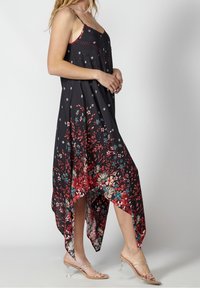 Femme portant une robe noire sans manches à motif floral rouge et turquoise avec un ourlet asymétrique, associée à des sandales à talons à brides transparentes.
