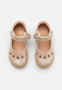 Beige patenttijäljitelmä nahkakengät, joissa on aukko-yksityiskohdat, pyöristetyt varpaat ja teksturoitu ruskea pohjallinen. Säädettävä hihna kultaisilla yksityiskohdilla.