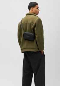 Schwarze Crossbody-Tasche mit Reißverschluss, getragen auf einer olivgrünen Fleecejacke. Verfügt über ein elegantes Design und einen verstellbaren Riemen.