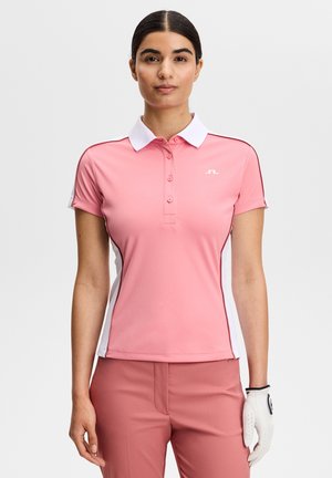 Kobieta ubrana w różową koszulkę golfową z białym kołnierzykiem i bocznymi panelami, do tego pasujące różowe spodnie oraz biała rękawica golfowa na lewej ręce.