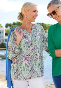Blusa leggera con motivo floreale nei toni del verde, rosa e blu; presenta un collo a V, maniche lunghe e dettagli con bottoni. Abbinata a uno strato esterno blu.