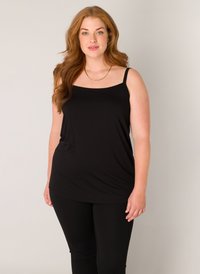 Vrouw met lang rood haar, gekleed in een zwart mouwloos topje en zwarte broek, staand tegen een effen lichte achtergrond.