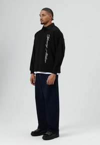 Zwart shirt met lange mouwen en een kraag, met witte scriptdetails, gecombineerd met donkerblauwe broek met brede pijpen en zwarte schoenen met roze veters.