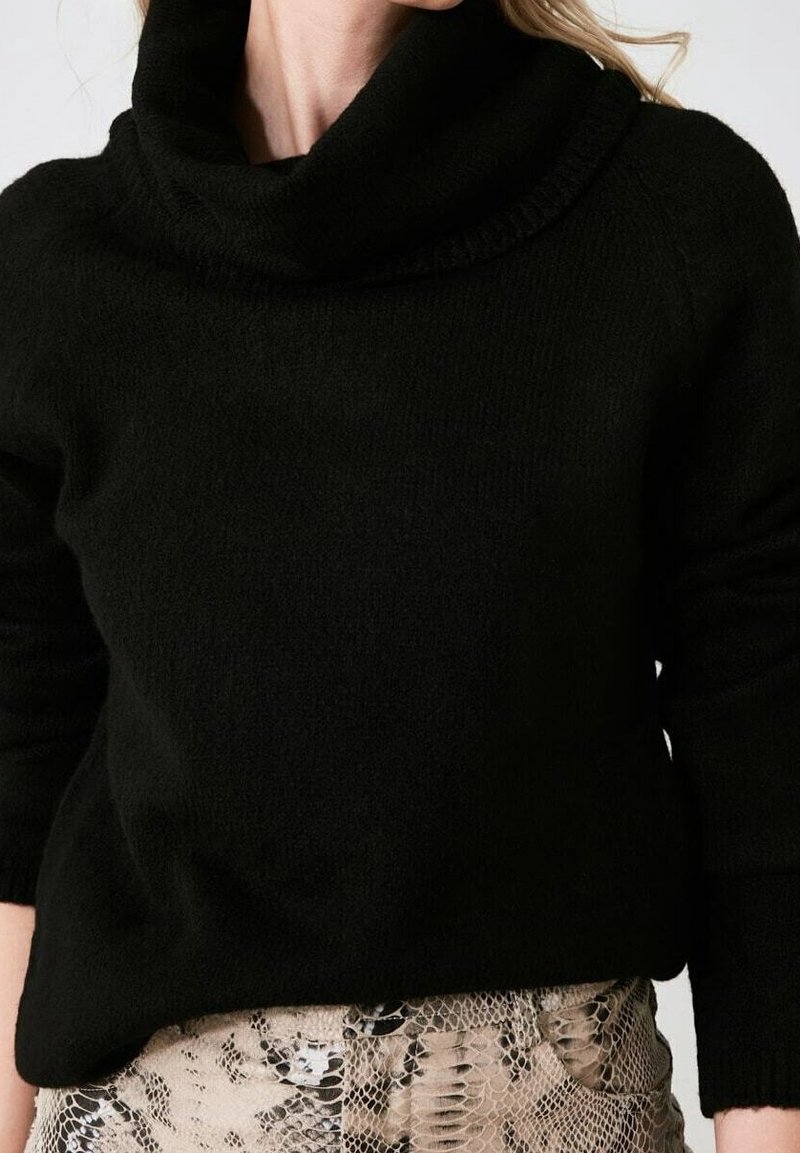Frau in einem schwarzen Rollkragenpullover, kombiniert mit beigen und schwarzen Schlangenleder-Musterhosen.