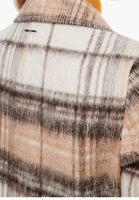 Chaqueta de punto de lana con un gran patrón de cuadros en crema, beige y marrón, con un acabado texturizado y una etiqueta de metal de la marca en la parte posterior.