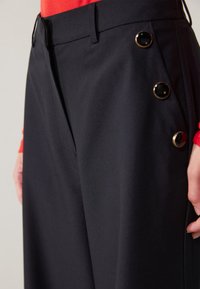 Black tailored trousers made of smooth fabric; features side pockets and three large, shiny black buttons as accents on one side.