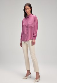 Camicia rosa con colletto e una tasca sul petto, abbinata a pantaloni slim-fit crema e scarpe con tacco di colore chiaro.