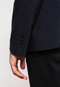 Handen i avslappnad position bredvid en marinblå kostymskjorta med fyra svarta knappar, över svarta byxor.