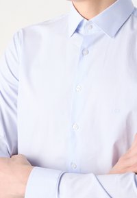 Camisa de botones a rayas azul claro y blanca, con un cuello clásico y botones blancos, que presenta un discreto bordado de logo en el pecho.