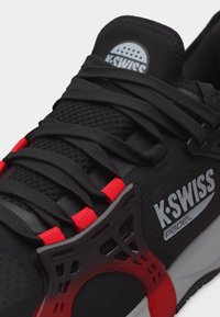 Μαύρο παπούτσι Padel της K-Swiss με μαύρα κορδόνια, κόκκινους βρόγχους κορδονιών και λογότυπο της μάρκας στη γλώσσα και στο πλάι.