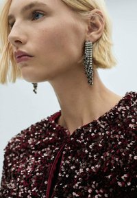 Giacca rossa con paillettes e texture lucida, dotata di un colletto alto e chiusura con zip, abbinata a orecchini pendenti in argento.