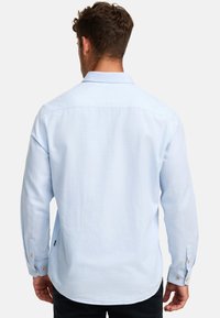 Uomo con capelli castani corti che indossa una camicia azzurro chiaro a maniche lunghe con bottoni, visto da dietro su uno sfondo semplice.