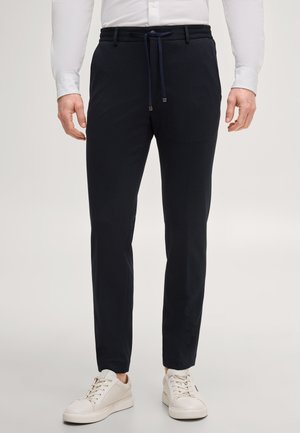 Mann trägt schwarze, schmal geschnittene Hose mit Tunnelzugbund und weiße Sneakers, steht vor einem schlichten hellen Hintergrund.