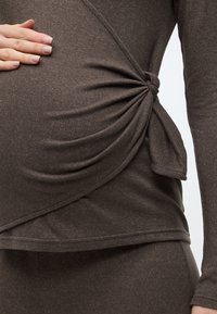 Καφέ τοπ εγκυμοσύνης από απαλό, ελαστικό ύφασμα. Λεπτομέρεια με κόμπο στο πλάι δημιουργεί εφέ draping, συμπληρώνοντας μια εφαρμοστή σιλουέτα.