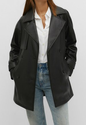 Femme portant une veste en cuir noire à double boutonnage sur une chemise blanche à boutons et un jean bleu clair, mains dans les poches de la veste.