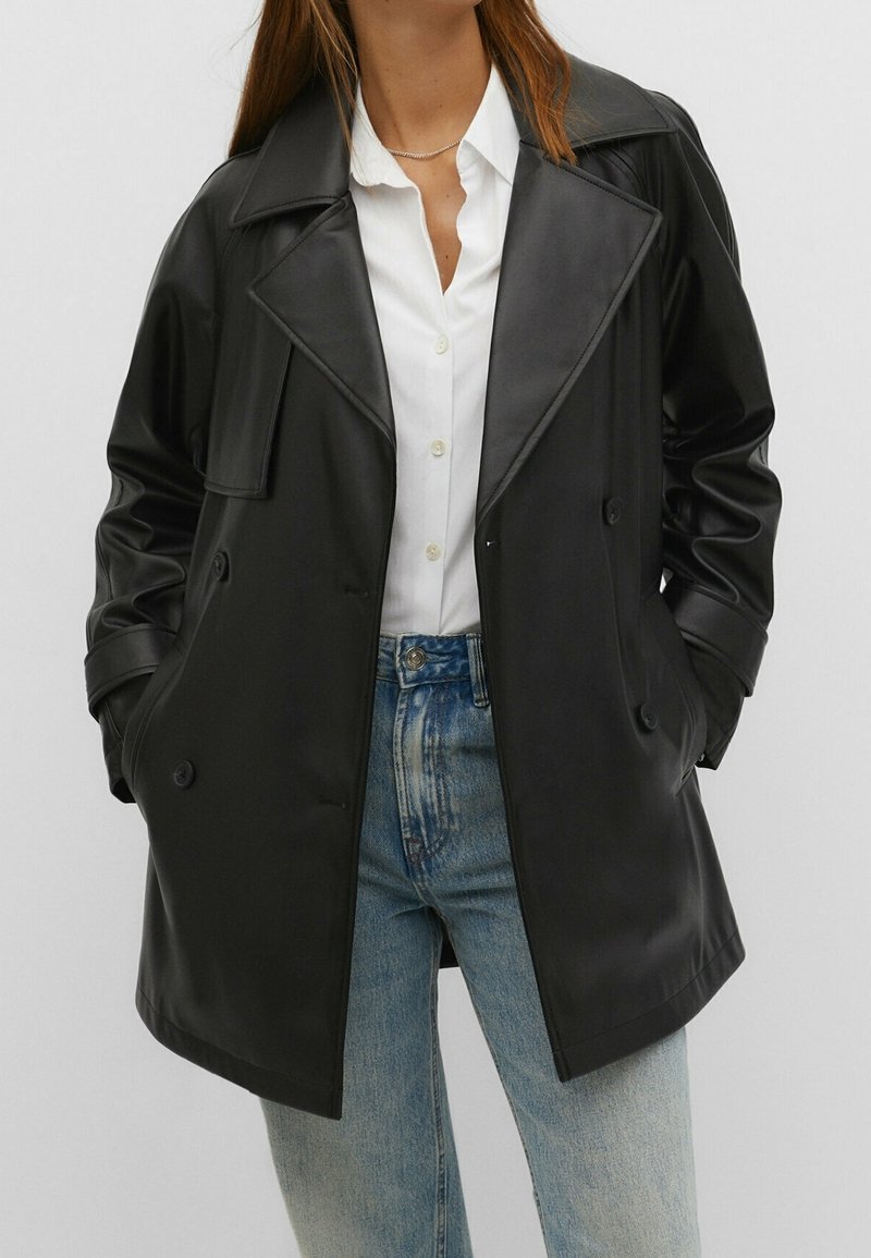 Femme portant une veste en cuir noire à double boutonnage sur une chemise blanche à boutons et un jean bleu clair, mains dans les poches de la veste.