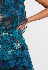 Vestido floral con tela superpuesta y transparente en tonos de azul verdoso y negro. Presenta patrones de mariposas y un dobladillo con volantes, mostrando texturas intrincadas.