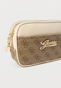 Pruuni mustriga kangast kott beežide äärtega, kahe tõmblukuga, kuldsete detailidega ja südamekujuline ripats. Suurelt esile tõstetud "Guess" logo.